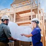 Two construction professionals wearing hard hats reviewing blueprints in front of a multi-story wooden building framework under construction on a sunny day