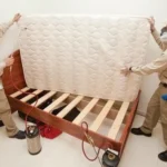 Two technicians lift a mattress while performing bed bug removal and treatment on a wooden bed frame