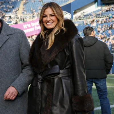 Smiling woman wearing a black leather coat with a wide fur collar and belted waist, standing on a sports stadium field with spectators in the background.