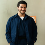 Smiling man wearing a navy blue cotton jacket and matching pants, standing against a light-colored wall with a relaxed pose